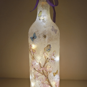 Standard wine bottle decorated with pink blossom tree and butterflies. Cork LED lights inside, switched on. Purple ribbon around the neck of the bottle.