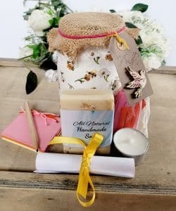 White jar with bee and flower design, hessian lid with card label with a dove on it. Contents shown outside of the jar, cream soap, small multicoloured scrapbook, short wooden pencil, tealight candle, small organza bag containing coloured paper quotes, white rolled up instruction leaflet tied with yellow ribbon.