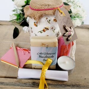 White jar with bee and flower design, hessian lid with card label with a dove on it. Contents shown outside of the jar, cream soap, small multicoloured scrapbook, short wooden pencil, tealight candle, small organza bag containing coloured paper quotes, white rolled up instruction leaflet tied with yellow ribbon.