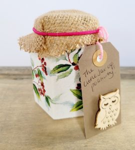 Hexagonal shaped jar painted white ith a green and red berry leaf design Hessian lid with brown tag. Tag states jar of positivity with a light beige owl on the bottom